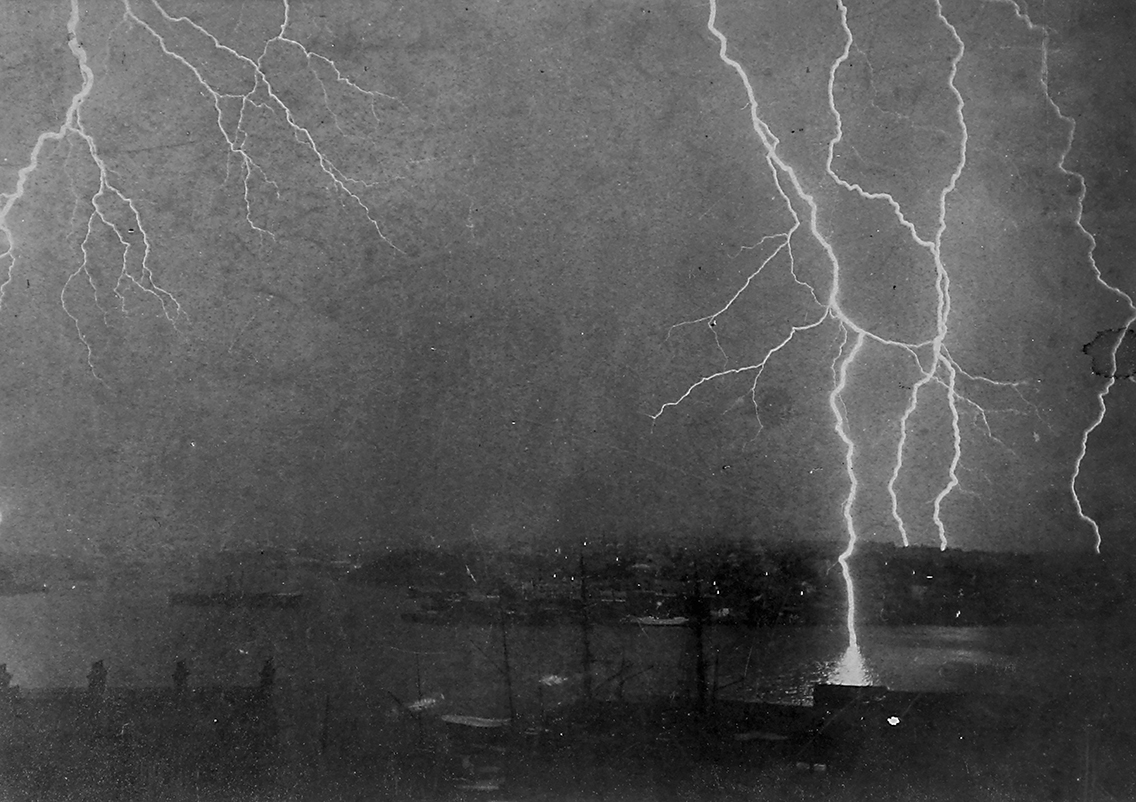Black and white photograph depicting a lightning strike at night. The top two-thirds of the image is filled by sky with multiple, branching bolts of lightning. In the foreground can faintly be seen various buildings and a body of water beyond.